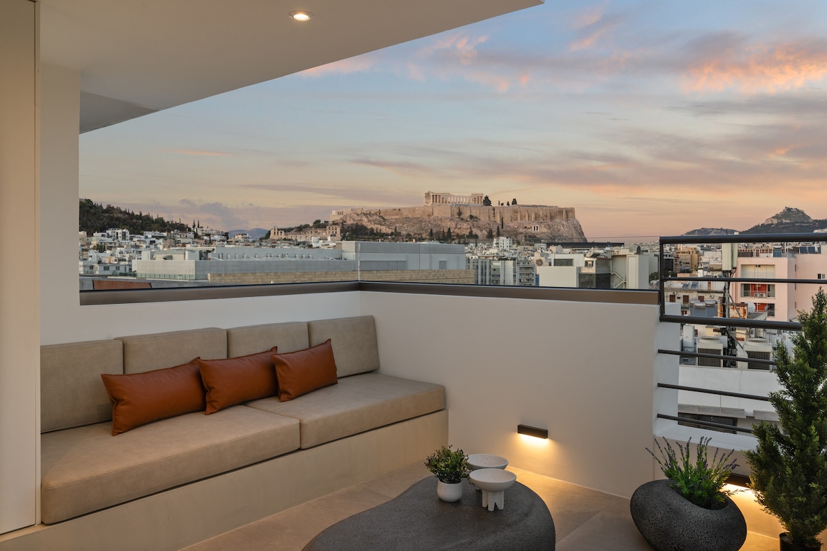 A spacious terrace features modern seating with earthy tones, overlooking the Acropolis. Potted plants add greenery, while soft lighting enhances the ambiance. The expansive view captures the historical site against a colorful sky, creating a serene outdoor retreat.
