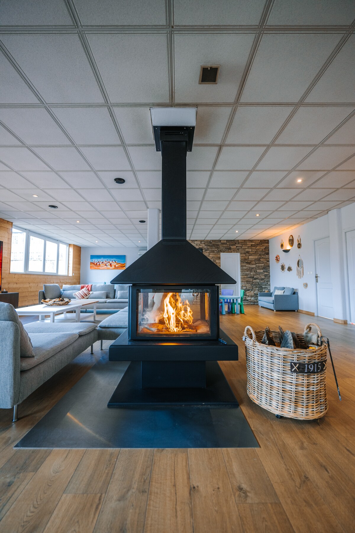 A central fireplace with a modern design is surrounded by elegant seating arrangements in a spacious living area. Natural light enters through large windows, while a wooden floor adds warmth. A woven basket for firewood is positioned nearby, enhancing the overall functionality of the space.