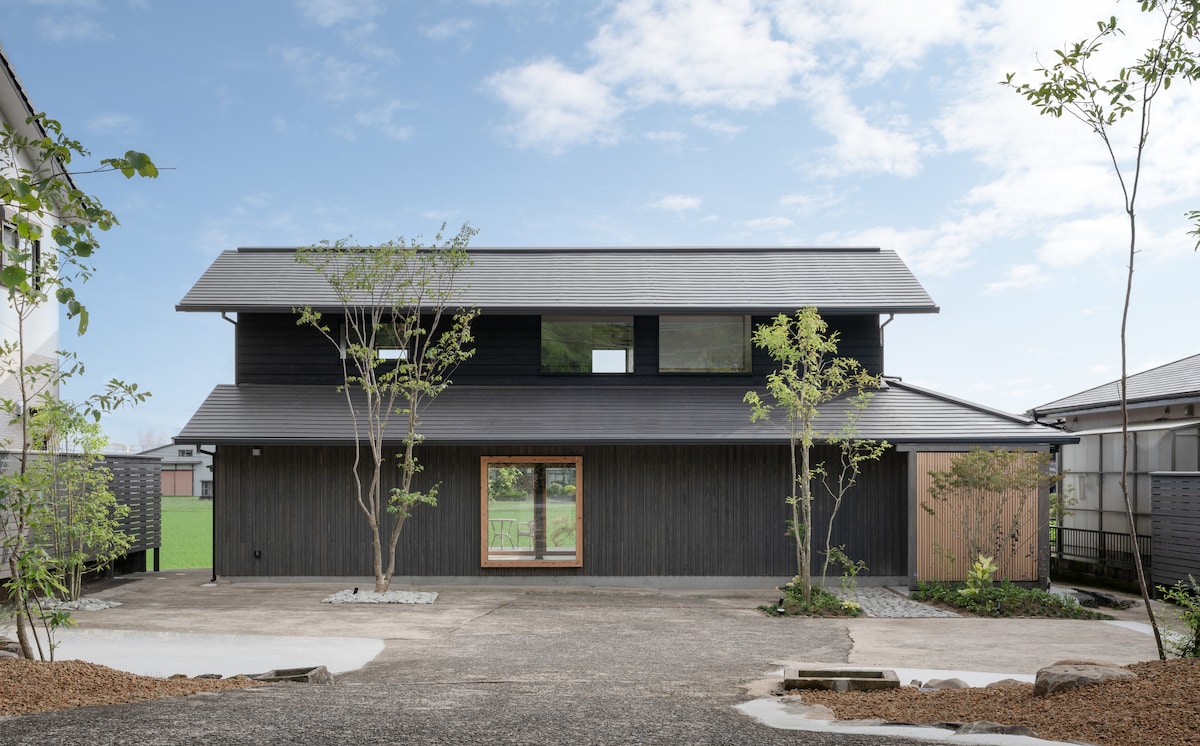 A traditional wooden house with a modern black exterior is set against a clear blue sky. The front features a central large window framed by minimal landscaping, while symmetrical trees and gravel pathways enhance the inviting entrance.