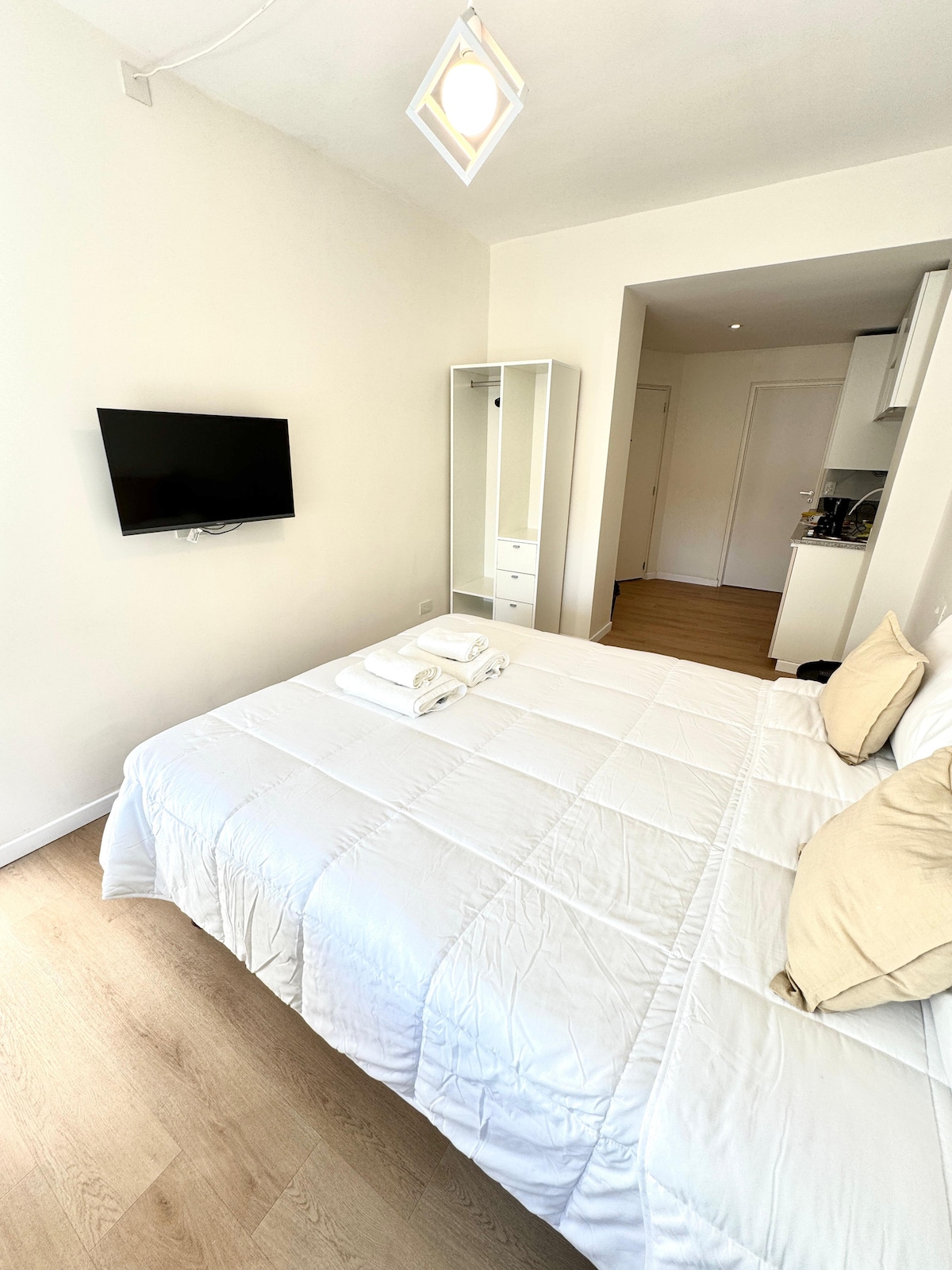 A well-lit sleeping area features a spacious bed with white bedding and decorative pillows. A flat-screen television mounted on the wall is visible. In the background, a modern kitchen area is partially seen, complemented by a wardrobe with mirrors enhancing the sense of space.