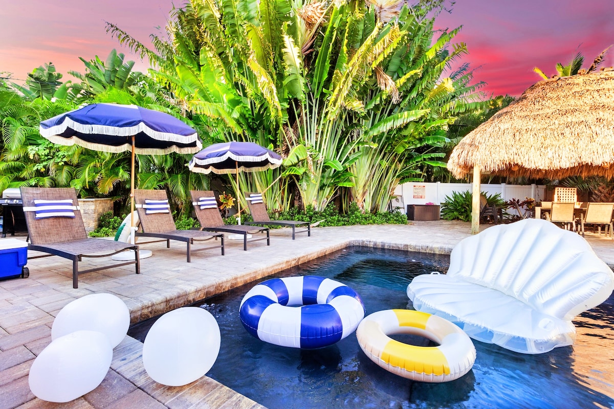 The backyard area features a salt-heated pool surrounded by tropical foliage. Lounge chairs with navy umbrellas are positioned nearby, along with various inflatable pool toys, creating a relaxing outdoor space for family entertainment.