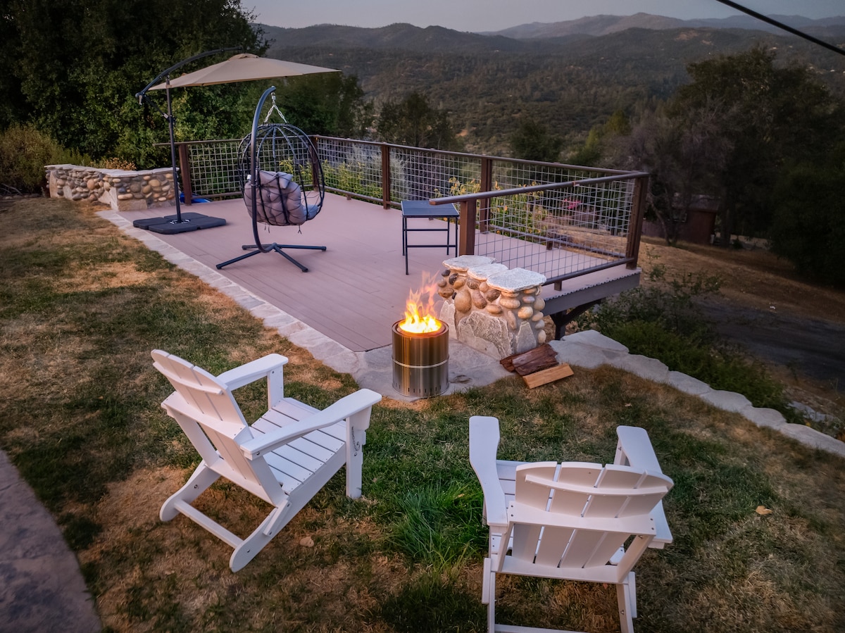 An outdoor deck features a fire pit surrounded by two white Adirondack chairs, providing a cozy gathering spot. A hanging chair adds a relaxed touch, overlooking expansive mountain views and a serene landscape. The scene captures a tranquil evening ambiance.