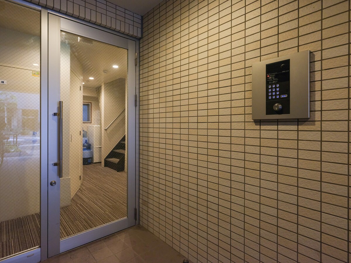 The entrance area features a glass door with a security intercom mounted on a tiled wall. Soft lighting creates a welcoming atmosphere. Visible stairs lead to upper levels, while the textured floor adds a subtle detail to the modern entryway.