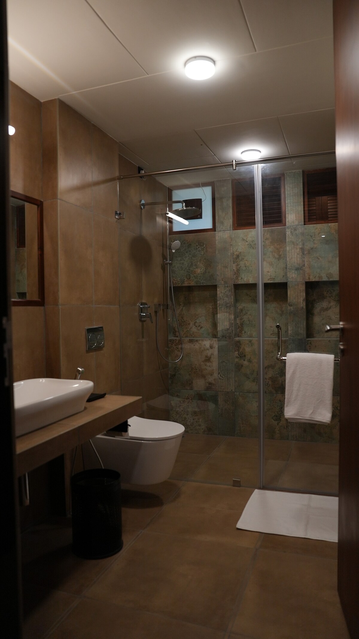 The bathroom features a spacious walk-in shower with a clear glass partition. A modern sink is placed on a wooden countertop, alongside a minimalistic toilet. Earthy tones create a calming atmosphere, with spotlights illuminating the textured wall tiles.