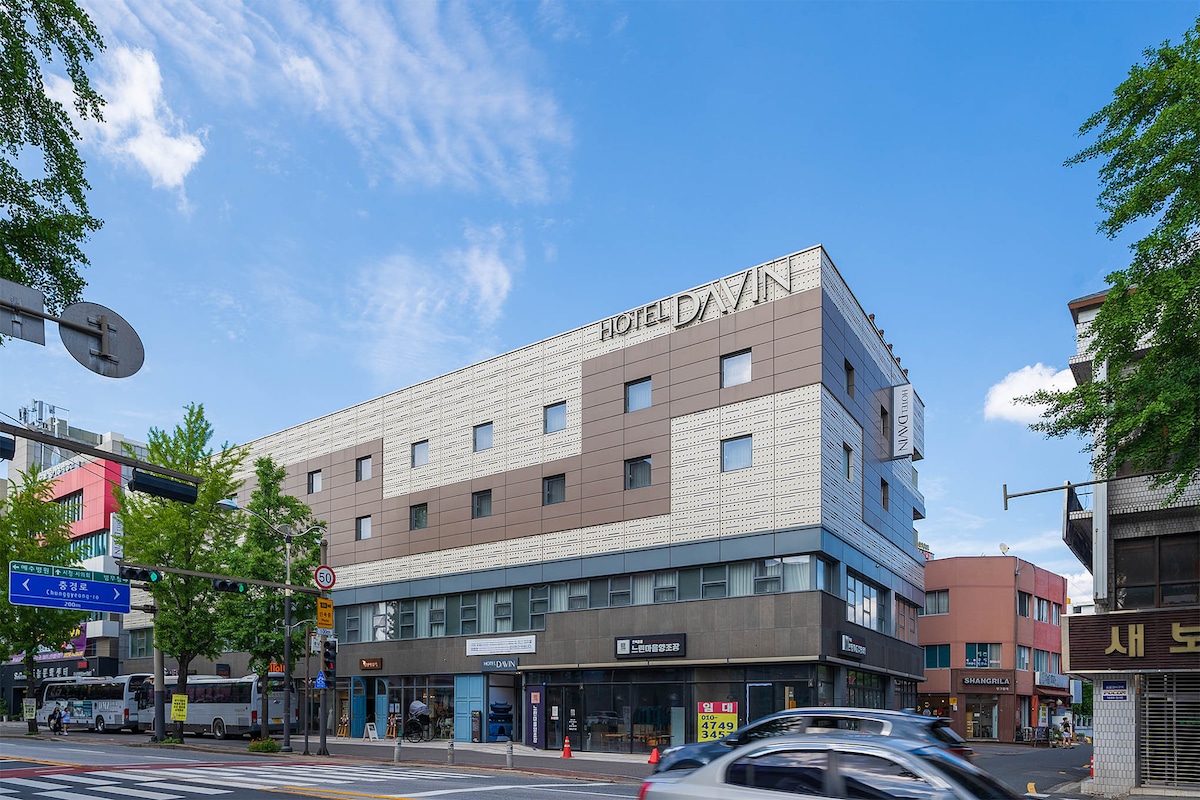 A modern hotel building is presented, featuring a sleek façade with a combination of light and dark panels. The structure is three stories high, and large windows are visible on each level. Surrounding greenery and nearby street signage contribute to the urban setting.