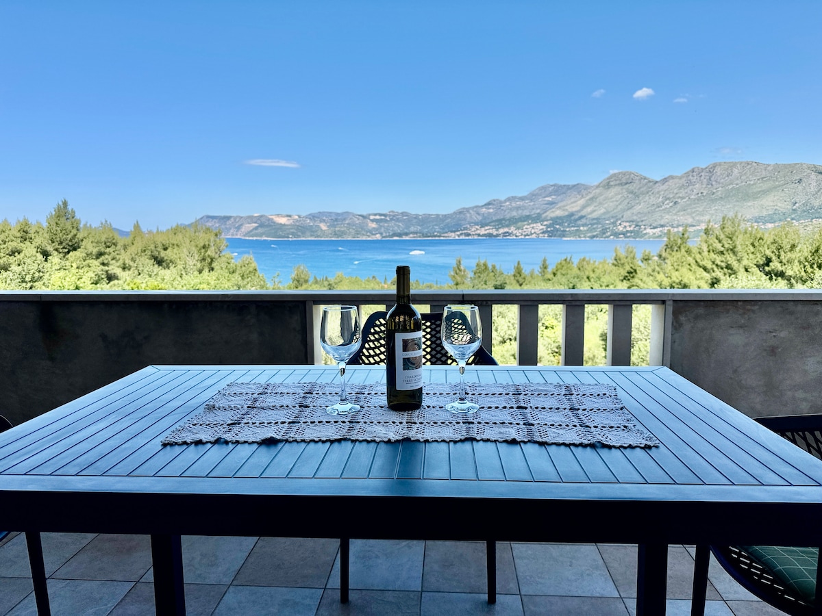 A dining table set for two stands on a balcony overlooking the sea. A bottle of wine and two glasses are positioned on a textured table runner, with lush greenery and distant hills framing the expansive water view under a clear blue sky.