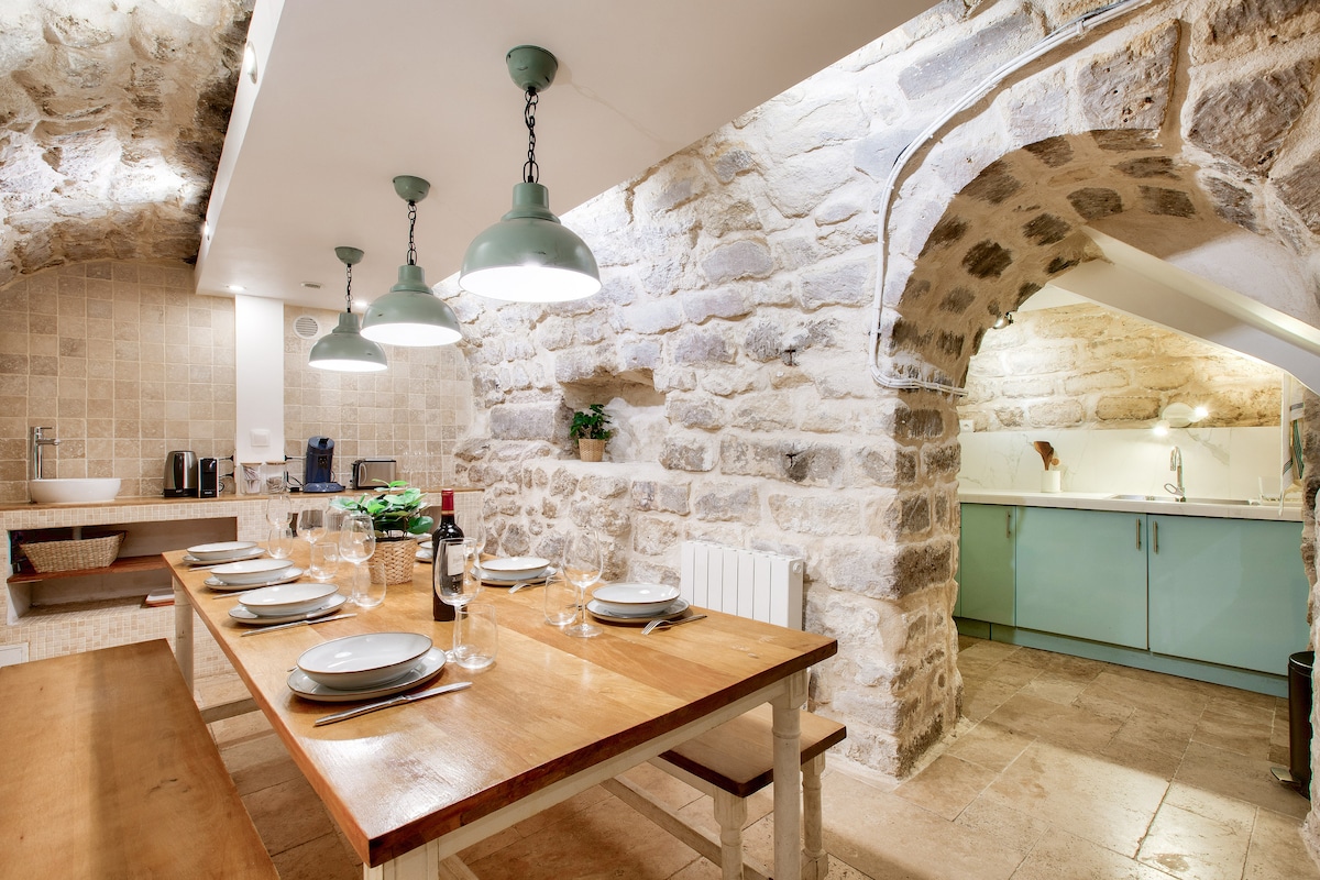 An inviting dining area features a rustic wooden table surrounded by chairs, with place settings prepared for a meal. Soft green pendant lights hang above, illuminating stone walls that add character to the space. A modern kitchenette with mint-green cabinetry is partially visible in the background.