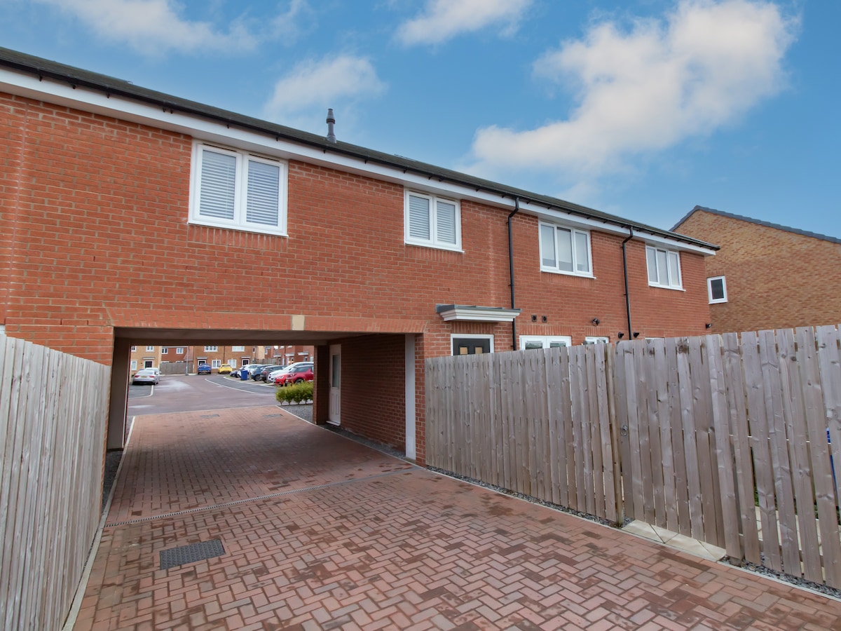 The exterior of the flat features a well-maintained brick façade with modern design elements. A designated parking area is visible beneath an archway, providing direct access to the building. Wooden fencing lines the property, ensuring privacy while allowing natural light to enter the space.