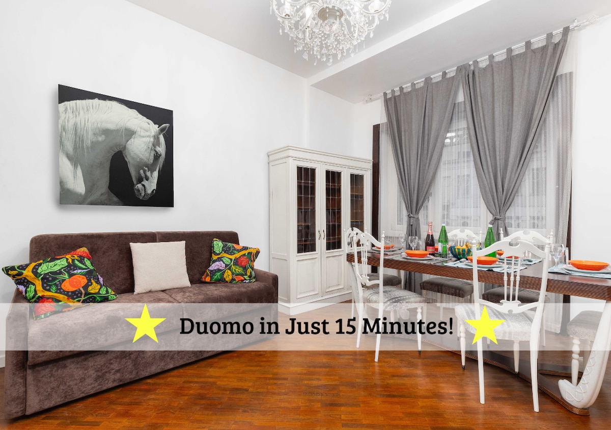 A bright dining area features a wooden table accompanied by white chairs, set for a meal with colorful plates. A spacious sofa in a rich brown hue is adorned with a mix of patterned cushions. Soft, natural light enters through sheer curtains, enhancing the room's inviting design.
