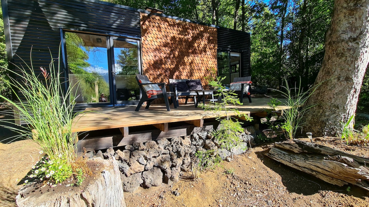 An outdoor deck made of wood is present, featuring comfortable seating with chairs positioned around a small table. Natural landscaping includes native greenery and stones, creating a harmonious connection with the surrounding forest.