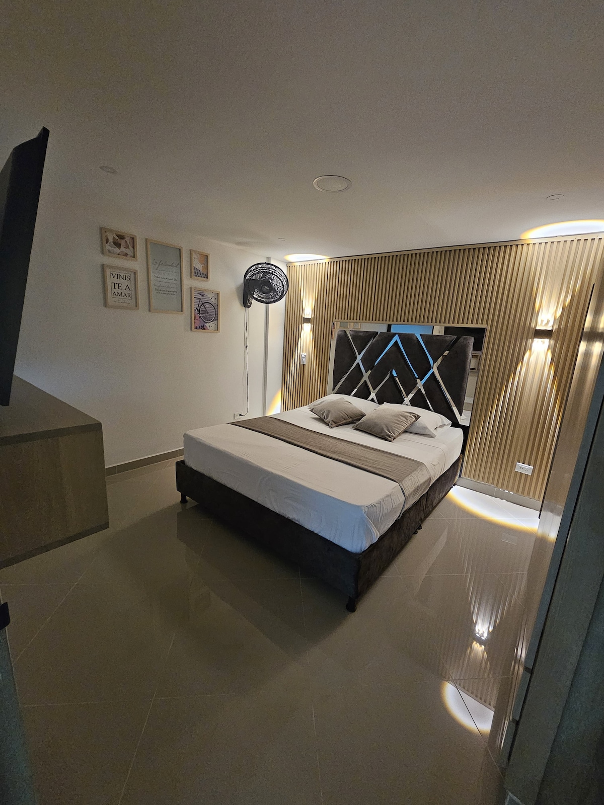 A modern sleeping area is presented with a spacious bed, adorned with neutral-toned bedding and decorative pillows. Soft lighting highlights the textured accent wall behind the headboard, while minimalist decor and a standing fan are visible, contributing to an uncluttered ambiance.