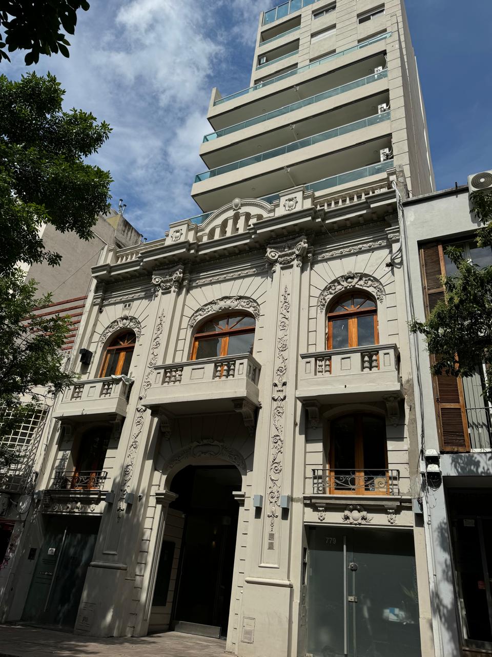 A historic building is showcased with ornate detailing on its facade. Large windows with wooden frames enhance the entrance, and greenery is visible at the base. A modern structure rises behind it, creating a juxtaposition of architectural styles.