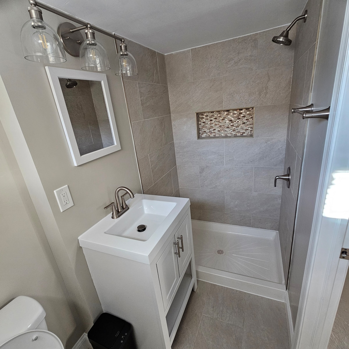A fully renovated bathroom is pictured, featuring a modern vanity with a white sink and sleek faucet. Soft lighting is provided by three glass pendant fixtures. The spacious shower area is tiled with neutral tones and includes a mosaic accent. A small mirror is mounted above the vanity.