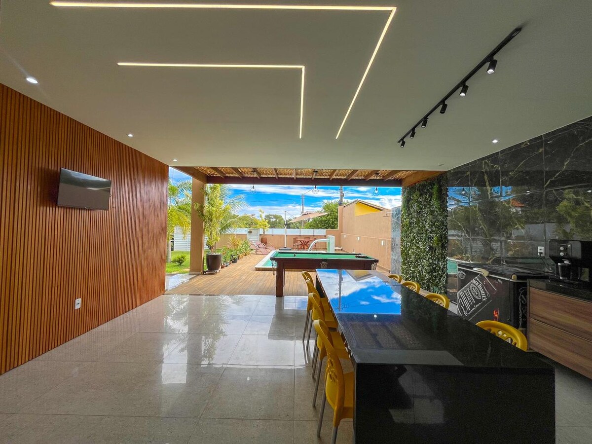 An open-plan area is highlighted, featuring a sleek dining table with yellow chairs, overlooking a pool and patio. Natural light floods in through expansive glass doors, creating a seamless transition to the outdoor space. Subtle ceiling lighting accentuates the modern design elements.