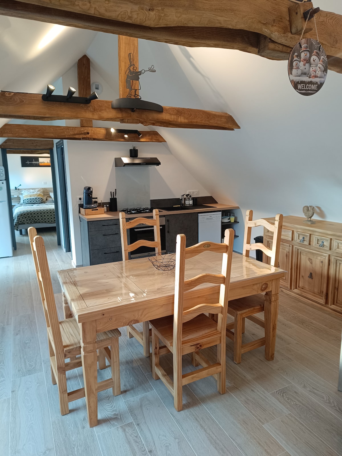 A cozy dining area is furnished with a wooden table and six chairs, complemented by a sideboard. Beams of natural wood highlight the sloped ceiling. The kitchen features a stove and modern amenities, creating a functional yet inviting space.