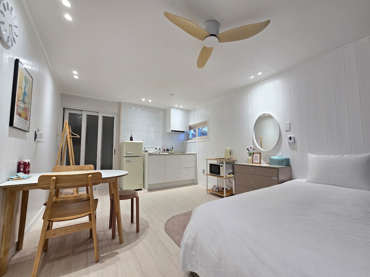 A bright and airy living space is presented featuring a spacious bed and minimalist décor. Natural light fills the area, highlighting a dining table and chairs, along with a kitchenette equipped with appliances. A large mirror reflects the cozy atmosphere while a circular clock adds functionality.