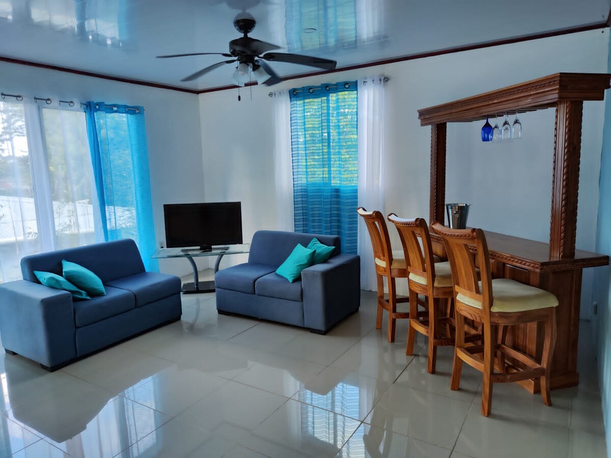 A well-lit living area features two blue sofas adorned with turquoise cushions. A flat-screen television is set on a glass stand opposite the seating. Behind, a wooden bar with high chairs is accented by three hanging glass light fixtures. Bright blue curtains frame the windows.