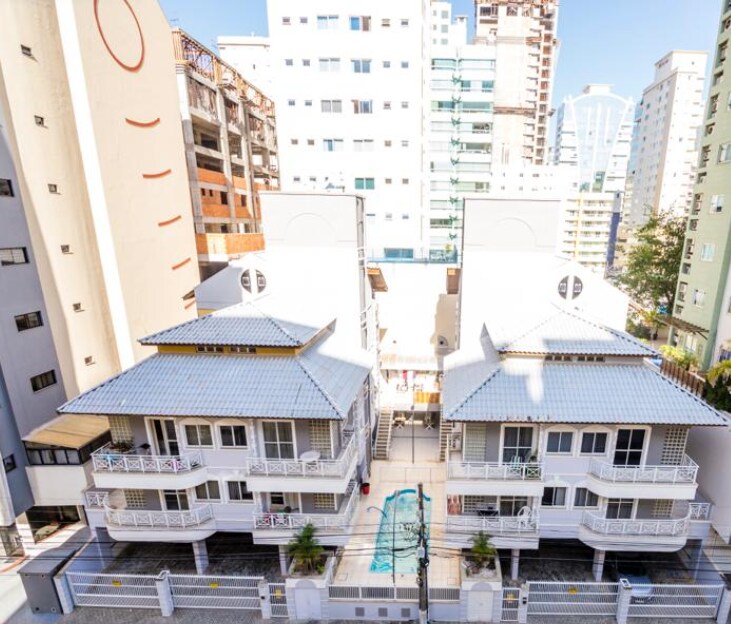 A view from above captures two adjacent buildings with tiled roofs. A central courtyard features a pool surrounded by palm trees, providing a focal point for outdoor activities. Nearby, urban structures rise, illustrating the blend of nature and city life.