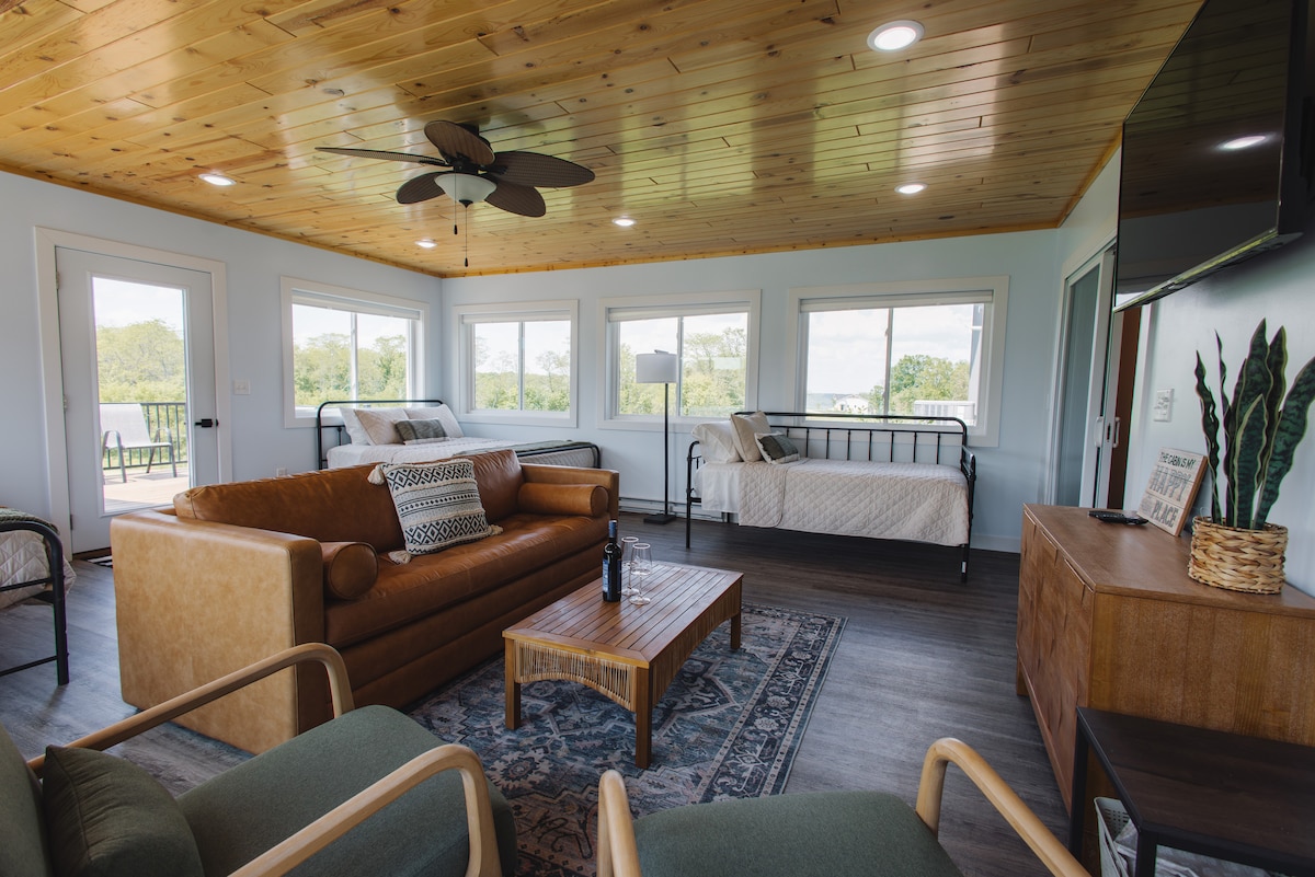 A spacious living area is highlighted by large windows offering natural light and views of the outdoors. A comfortable brown sofa is centered in the room, accompanied by two green chairs. A wooden coffee table rests in front of the sofa, with a daybed positioned near the window.