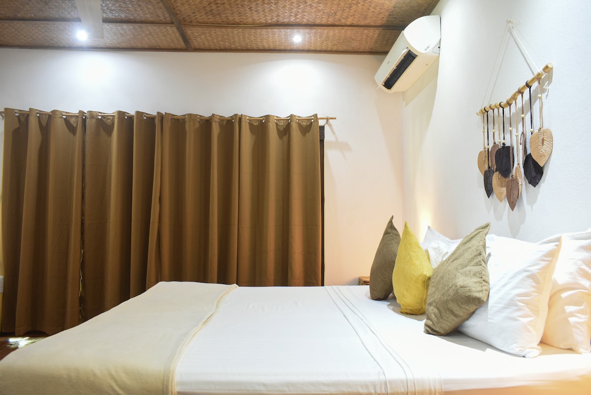 A spacious bed is situated against a textured wall, layered with neutral linens and accented by colorful throw pillows. Soft lighting creates a calming ambiance, while curtains frame the window, enhancing privacy. An air conditioning unit is mounted above for comfort.