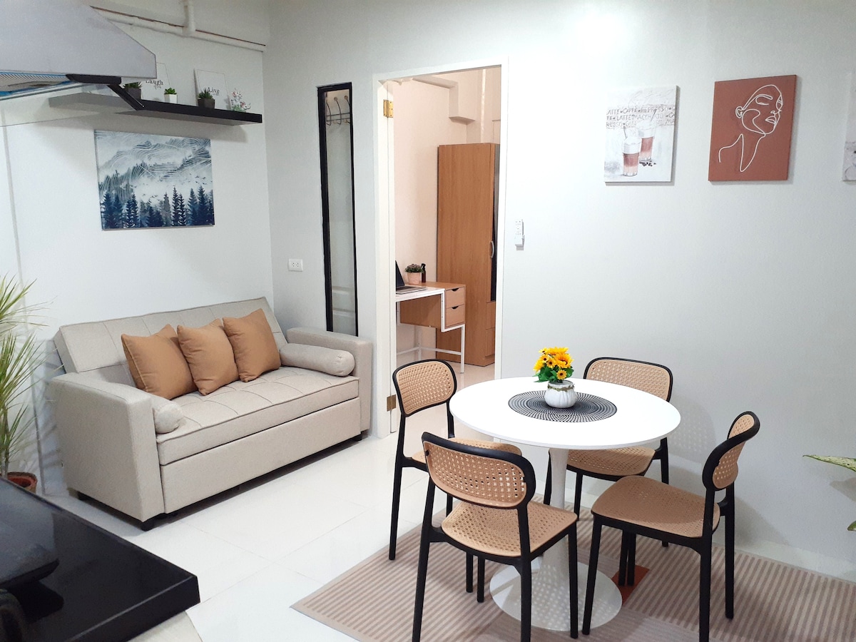An inviting dining area is furnished with a round table and four chairs, complemented by a cheerful centerpiece of flowers. A cozy sofa with decorative cushions is positioned nearby, with artwork adorning the walls and a doorway leading to a separate room.