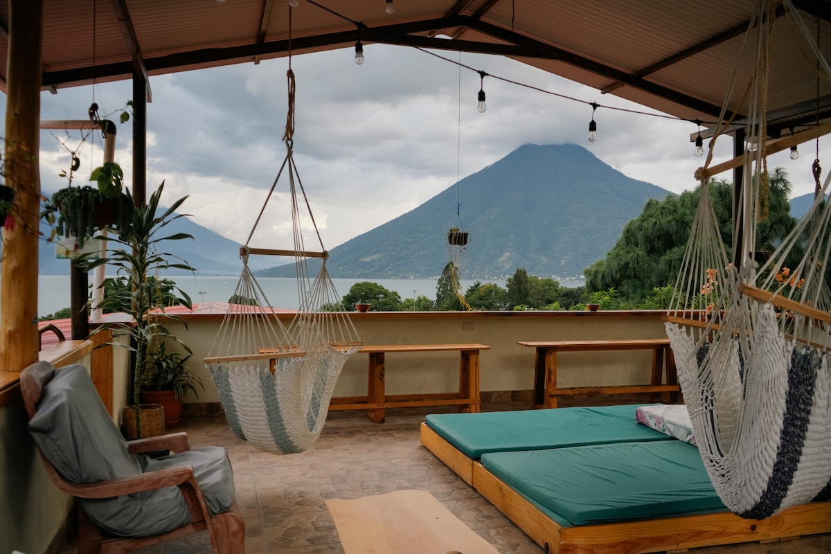 A spacious rooftop area features hanging hammocks and cushioned seating, with a stunning view of the surrounding mountains and a nearby lake. Natural wood benches provide additional seating, while the soft lighting creates a serene atmosphere, inviting relaxation and enjoyment of the landscape.