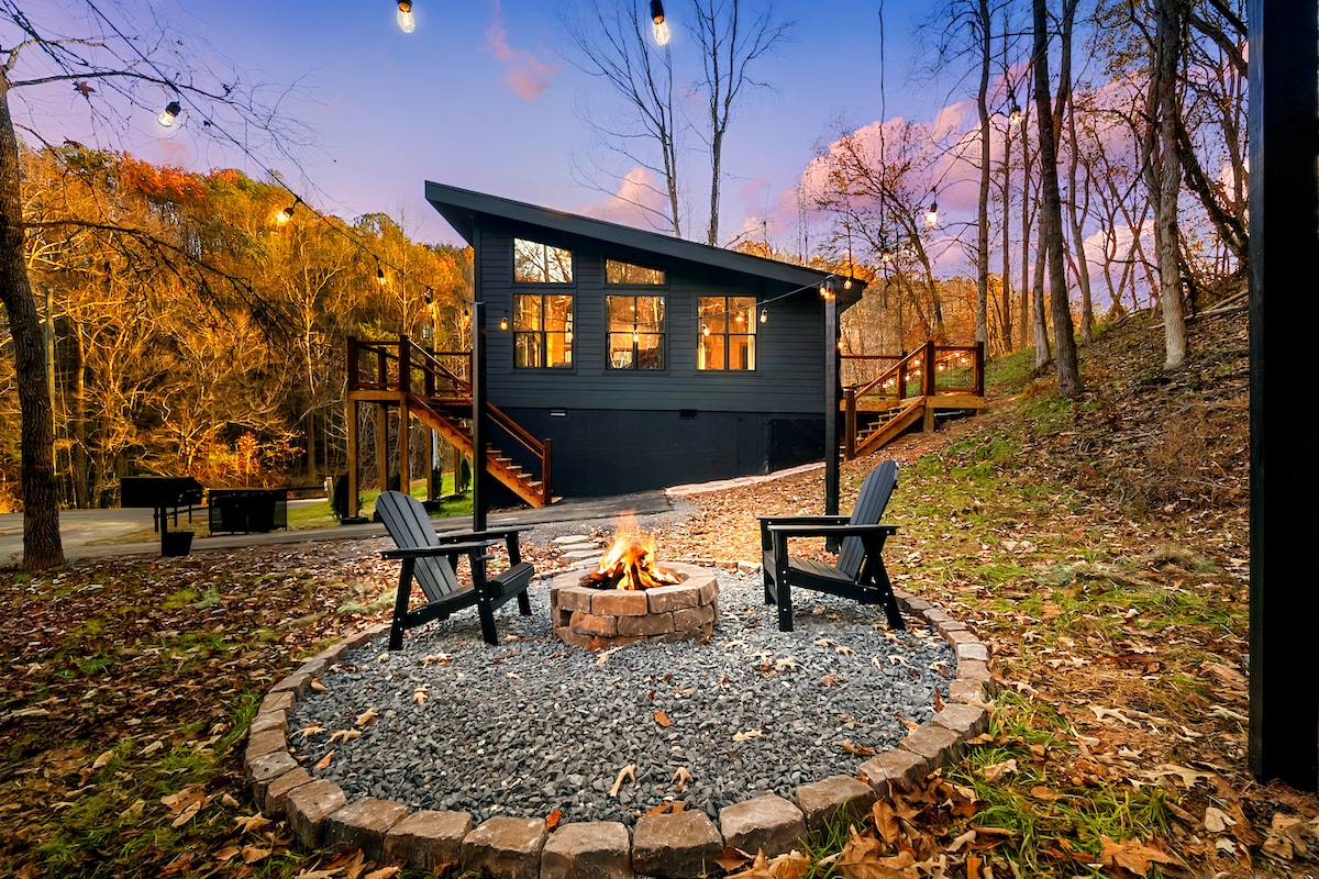 A custom fire pit surrounded by gravel is flanked by two Adirondack chairs. The modern home, featuring large windows, is positioned among trees with autumn foliage. Warm lights are strung above the outdoor area, creating a cozy ambiance as the sun sets.
