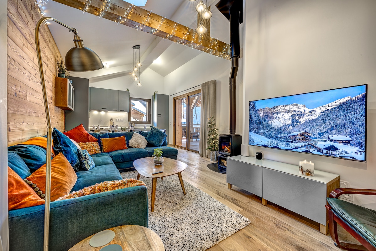 The open-plan living room features a large L-shaped sofa adorned with colorful cushions. A wooden coffee table rests on a soft area rug, while a smart TV is mounted on the wall. Natural light floods through patio doors, illuminating the cozy wood stove.
