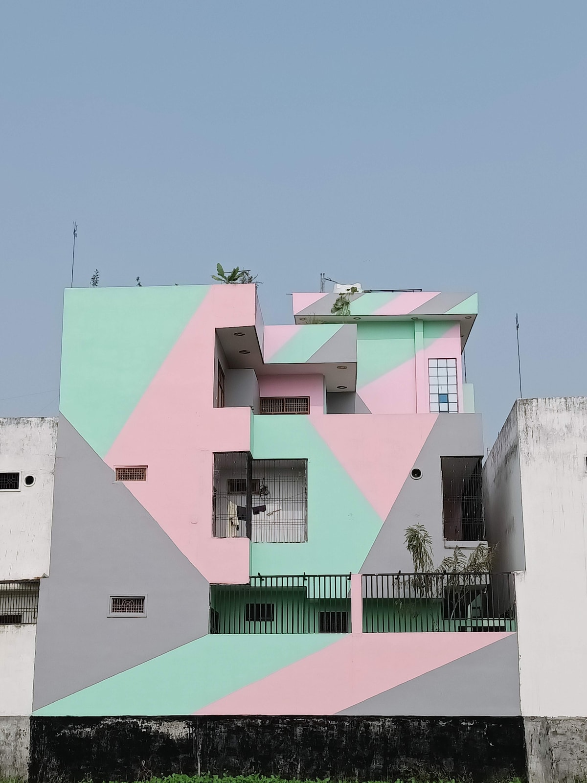 A creatively designed building showcases bold geometric patterns in pastel shades of pink and green. The structure features multiple levels, with terraces enhanced by greenery. Clear sky serves as a backdrop, highlighting the building's unique architectural style.