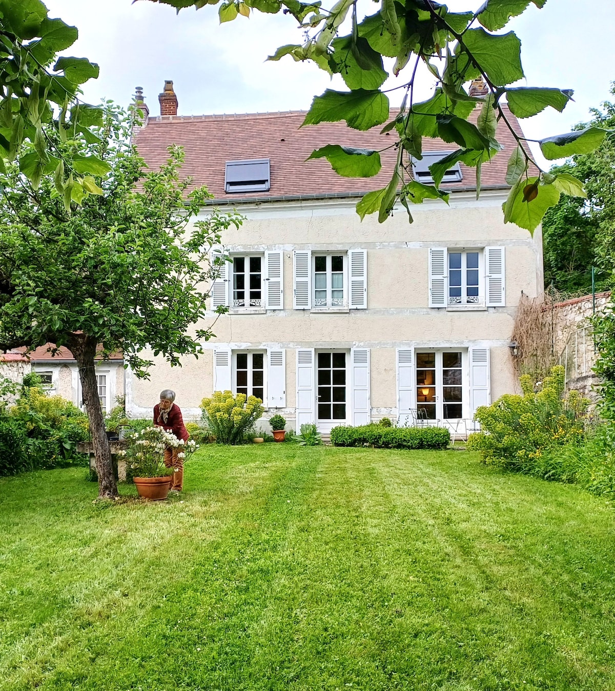 The exterior of a charming house is visible, showcasing a light-colored facade with multiple windows and traditional shutters. A neatly landscaped garden features green grass and potted plants. The area is shaded by a tree, creating a serene outdoor space.