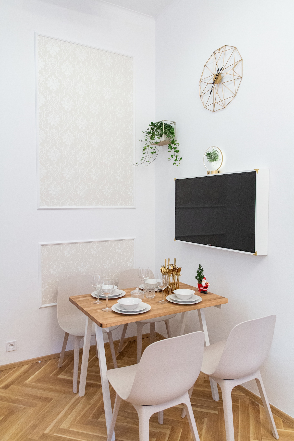 A dining area is set for four, featuring a light wood table with plates, glasses, and cutlery arranged neatly. The wall is adorned with a decorative clock and greenery, creating a minimalistic aesthetic. Light wooden flooring contrasts with the white walls.