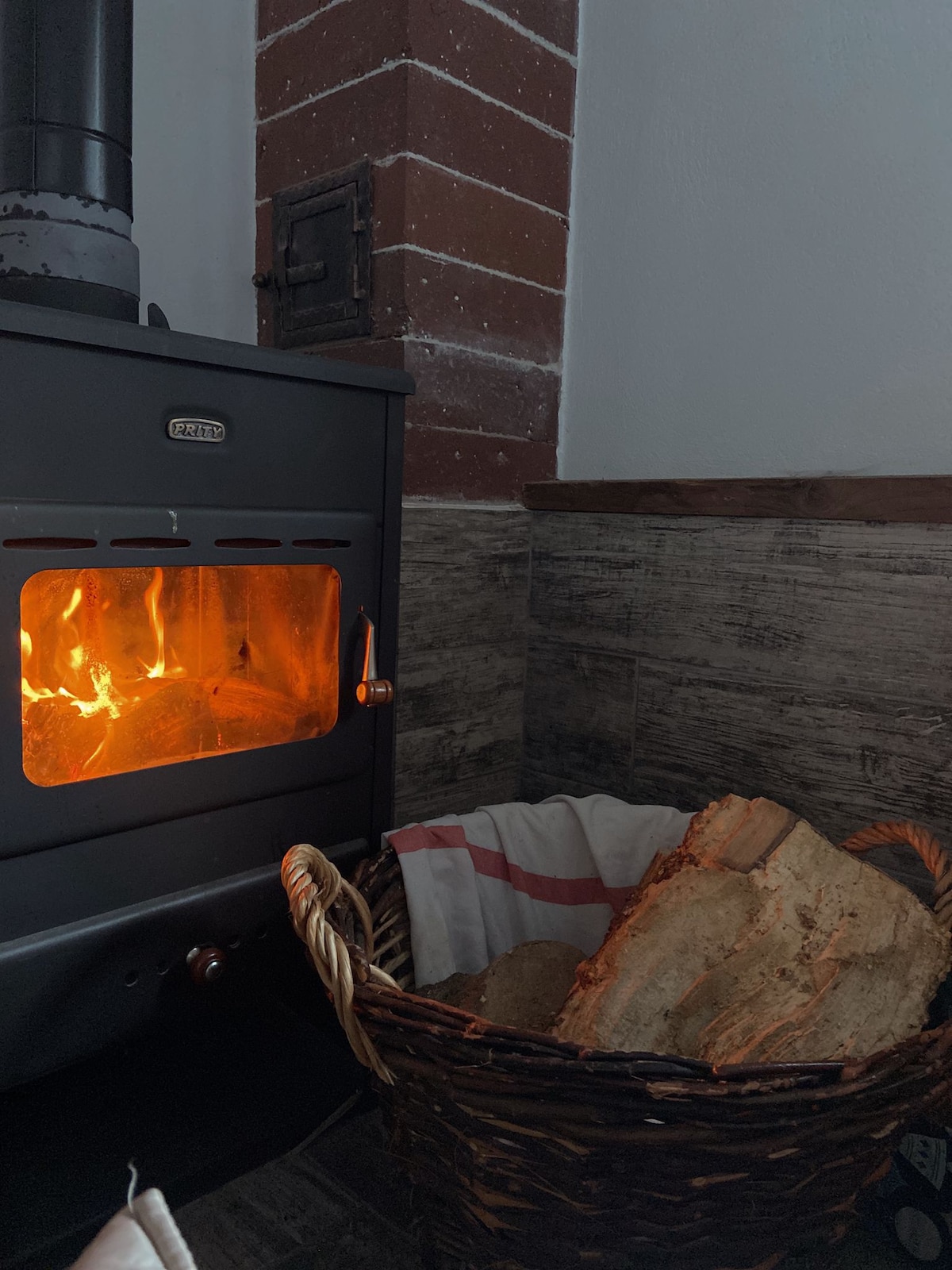 A wood-burning stove is positioned against a textured wall, featuring a glowing flame visible through its glass door. A woven basket beside the stove holds neatly stacked firewood, contributing to a warm and cozy ambiance.