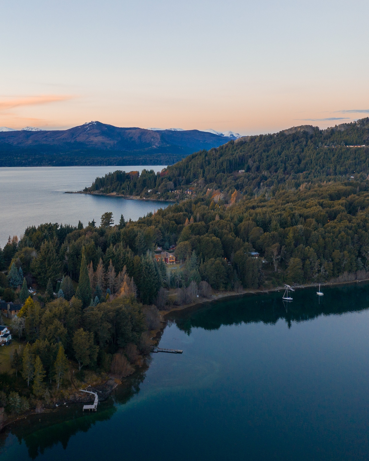 An aerial view captures the serene landscape surrounding Lake Nahuel Huapi. Lush green forests are interspersed with residential structures. Majestic mountains frame the horizon, while the calm water reflects the soft hues of the evening sky, creating a tranquil atmosphere.