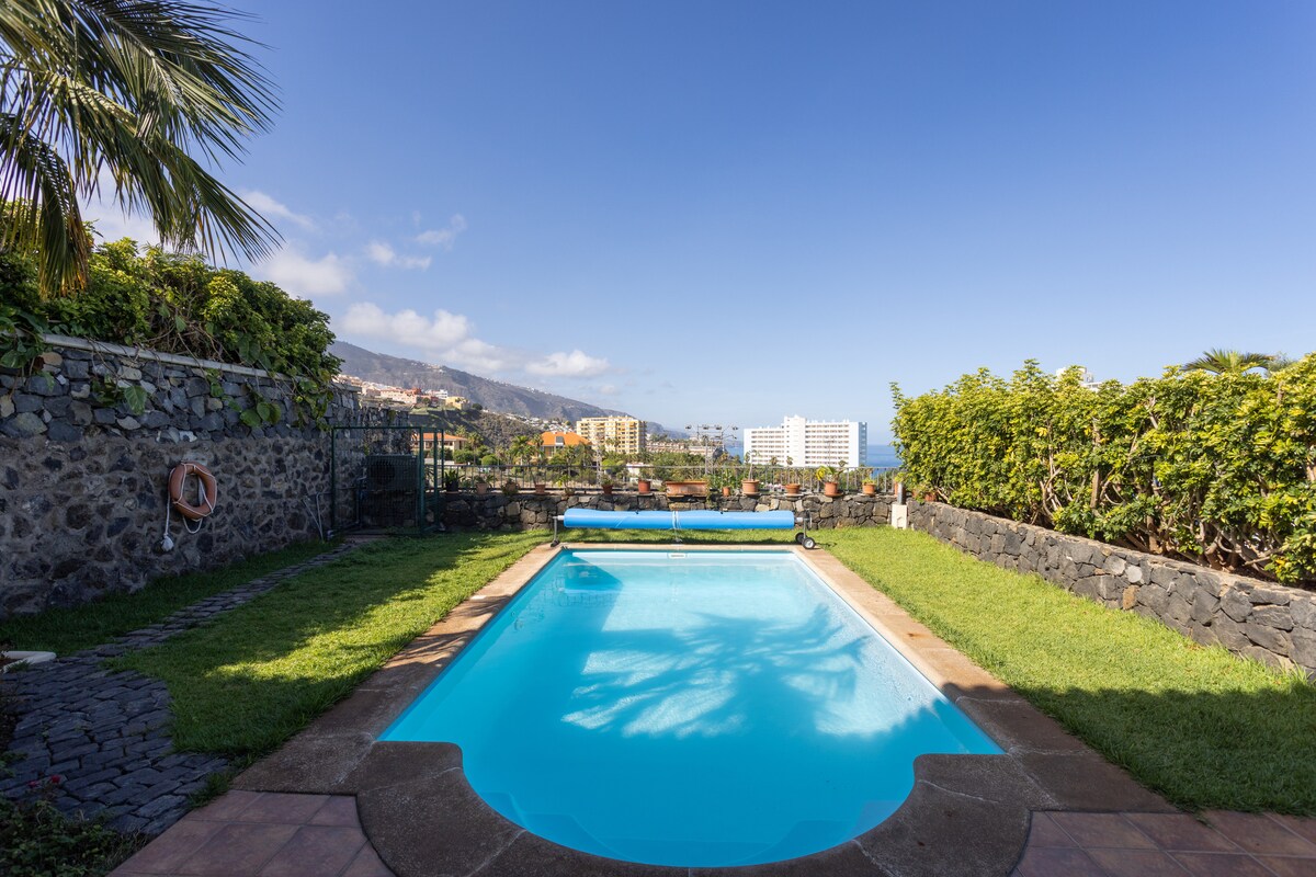 A heated swimming pool is situated in a well-maintained garden, offering views of the surrounding landscape and distant buildings. Lush greenery lines the pool area, with a stone wall providing privacy. Clear blue skies contribute to a serene and inviting atmosphere.