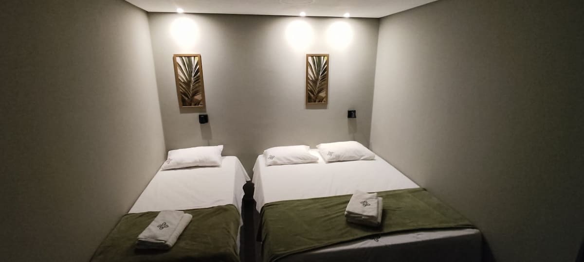 The room contains two spacious beds, each adorned with white linens and green throws. Light fixtures are positioned above, softly illuminating the space. Decorative wall pieces featuring leaf motifs add a touch of natural elegance.