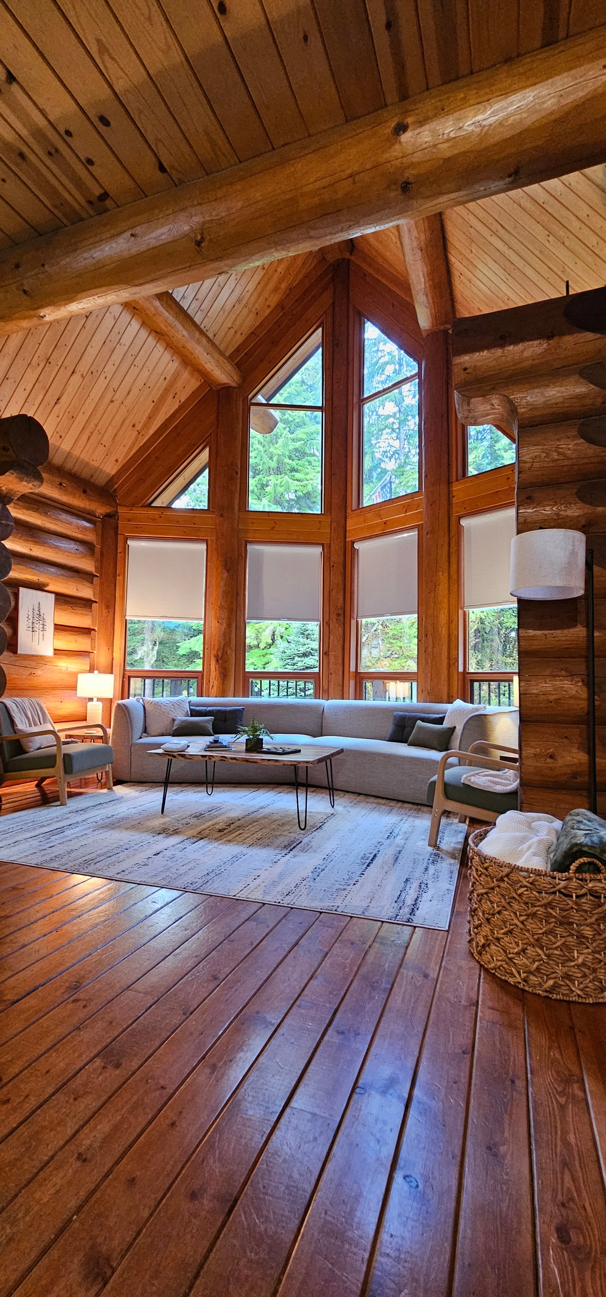 A spacious living area features large triangular windows that allow natural light to fill the space. The wooden interior showcases log accents and a polished floor. A comfortable couch and side tables are arranged around a central coffee table, creating an inviting setting for relaxation.