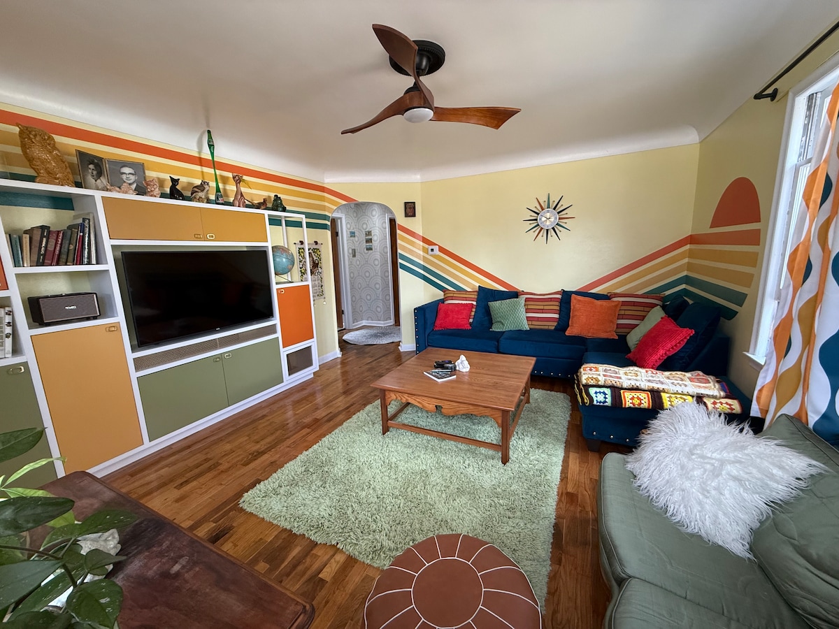 The living room features a mix of colorful furnishings and vintage decor. A cozy seating area includes a blue sectional sofa with patterned throw pillows, a wooden coffee table, and a plush area rug. Decorative wall designs add character, and a television is mounted on a stylish cabinet.