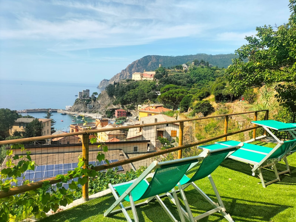 The image features a panoramic terrace overlooking a coastal village and the sea. Several sun loungers are arranged on a grassy area, inviting relaxation. The backdrop includes scenic cliffs and buildings descending toward the water, with distant shorelines visible under a clear sky.