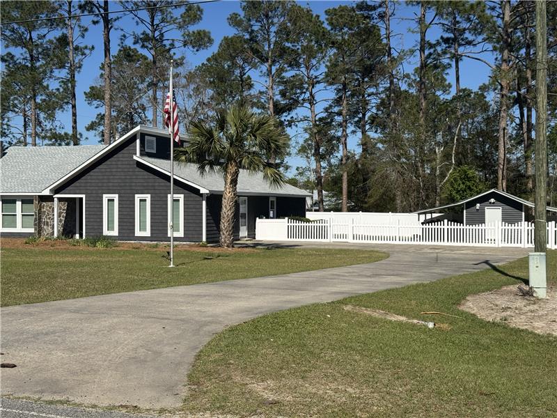 A charming home with a dark exterior is set against a backdrop of tall pine trees. A wide driveway leads to the house, with a white picket fence surrounding the property. An American flag is displayed in front, adding to the welcoming ambiance.