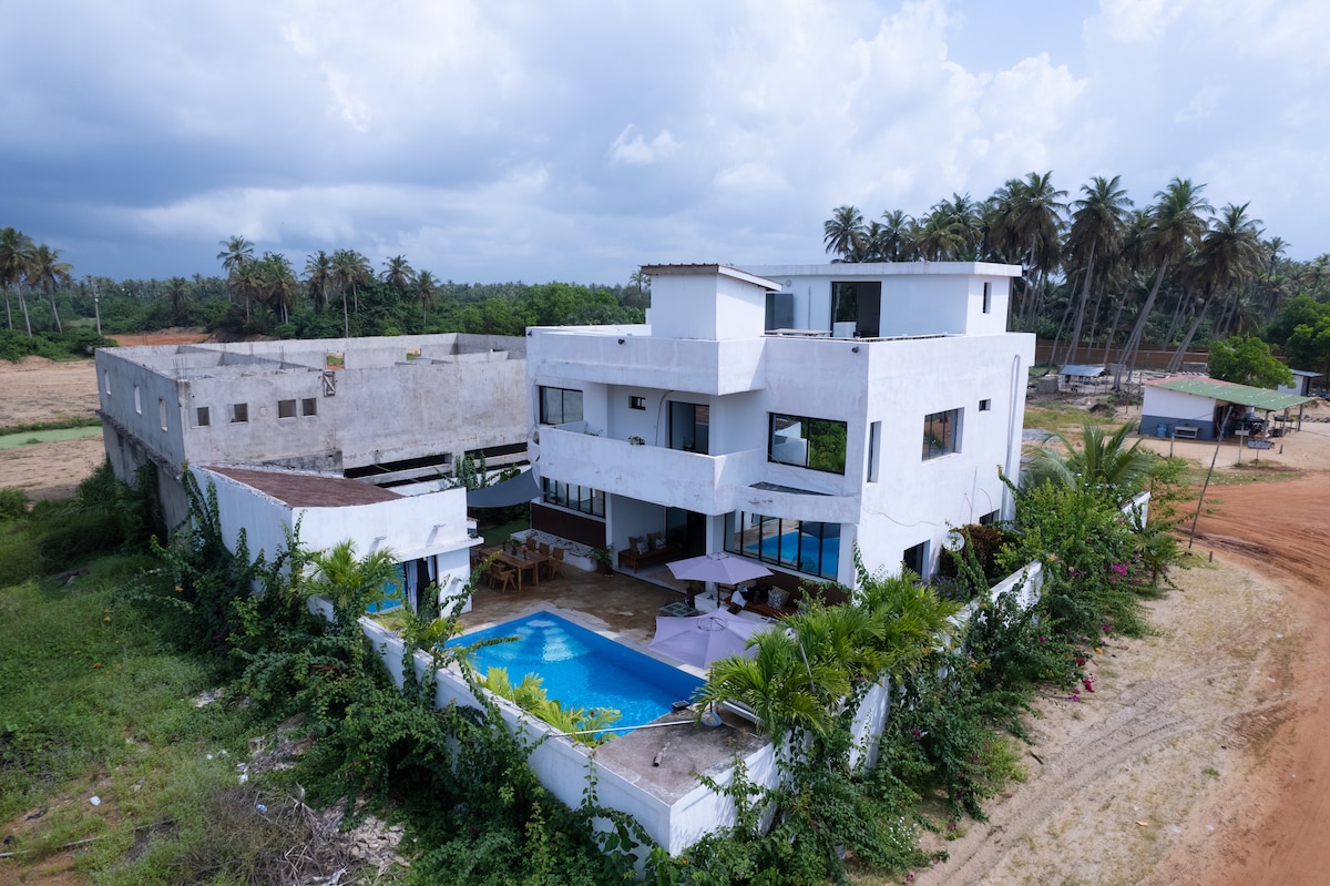 An aerial view of a modern villa showcases its clean white facade, surrounded by lush greenery and palm trees. A private pool features in the foreground, with seating areas visible nearby. The structure's design emphasizes spaciousness and light, with large windows on multiple levels.