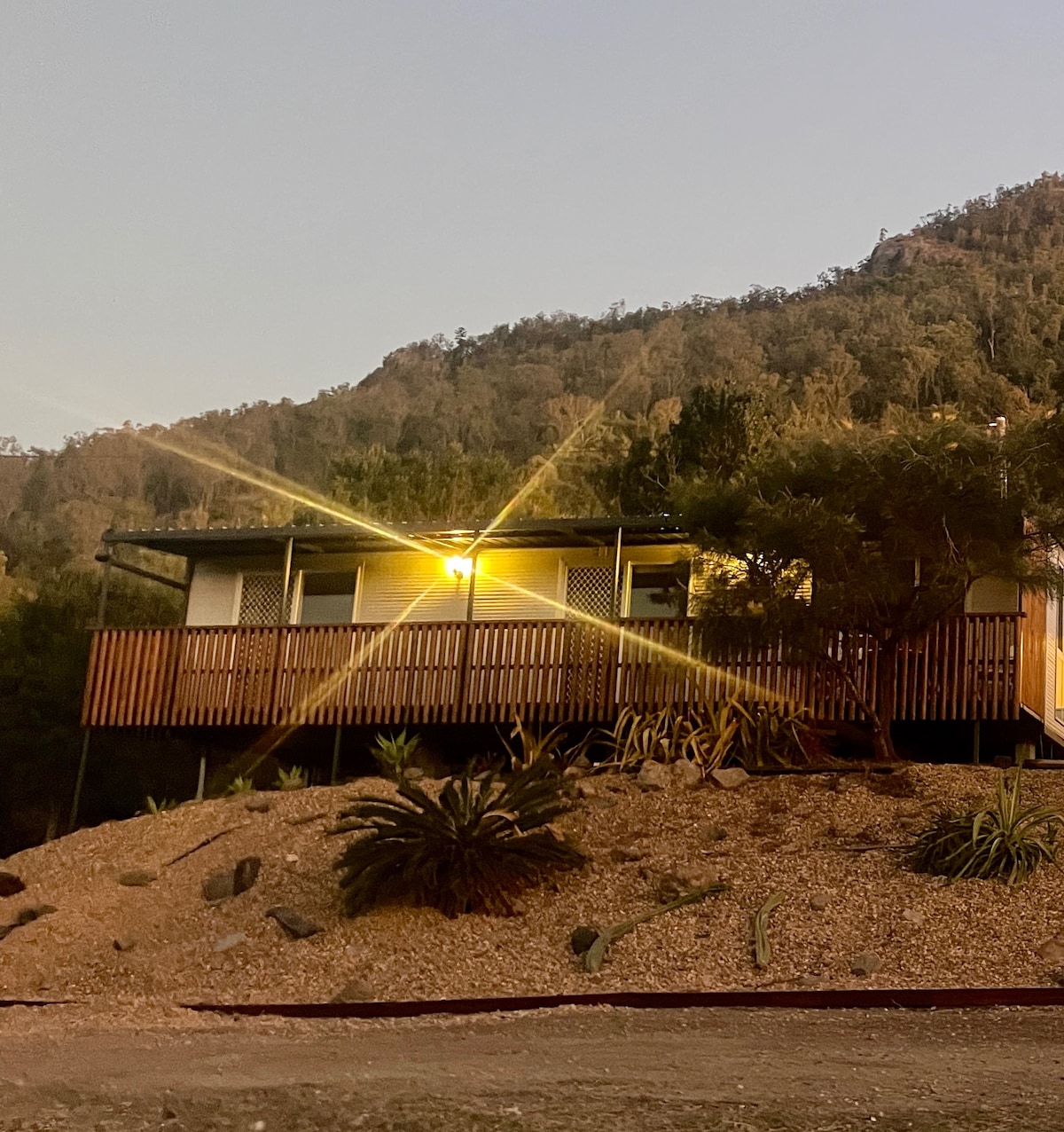 An evening view of the glasshouse is presented, showcasing its semi-attached structure elevated on a hillside. Soft lighting enhances the exterior, surrounded by native plants and gravel. The backdrop features gently sloping hills, creating a serene atmosphere as daylight fades.