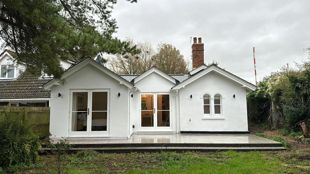 The exterior of the bungalow is depicted, showcasing a clean, white facade with large glass doors that lead to a welcoming entrance. The surrounding area features lush greenery and a small patio area. A chimney is visible, adding to the overall character of the property.