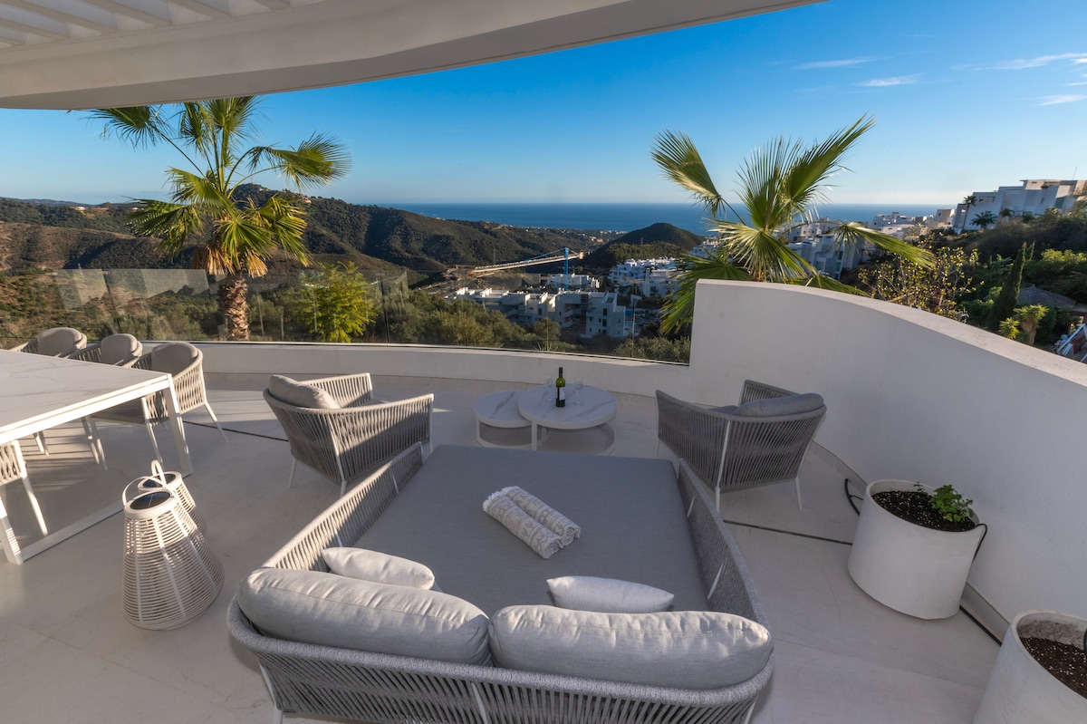 A terrace features modern outdoor furniture, including a spacious seating area with plush cushions. A view of rolling hills and the sea is visible in the background, framed by palm trees and greenery. A small table sits at the center, inviting relaxation.
