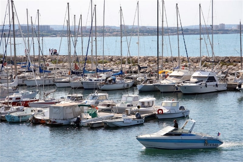 A marina is filled with various boats, both motorized and sailboats, floating gently on calm waters. The distant shoreline is visible, lined with low buildings under a clear sky. A group of people can be seen on the pier, enjoying the serene maritime environment.