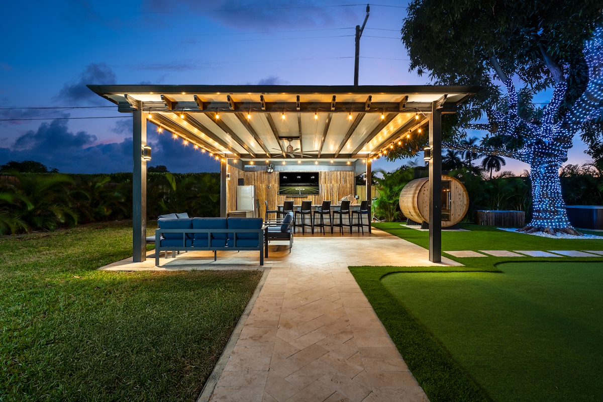 An outdoor entertainment space is illuminated softly by string lights, featuring a covered area with seating and a dining table. The grassy surroundings and a large tree, adorned with lights, create a serene atmosphere for gatherings as dusk approaches.