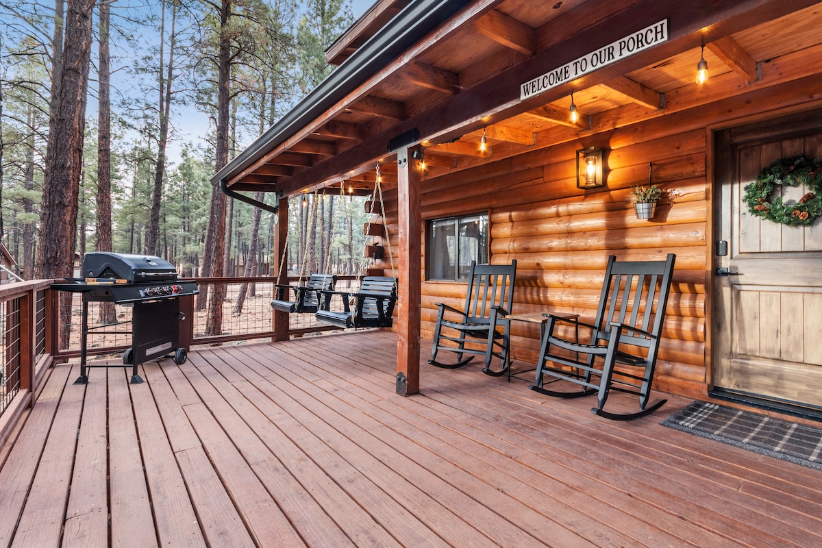 A spacious outdoor deck features wooden rocking chairs and a BBQ grill, inviting relaxation among the surrounding pines. Soft lighting enhances the welcoming atmosphere, while a sign reads 'Welcome to Our Porch' near the cabin entrance.
