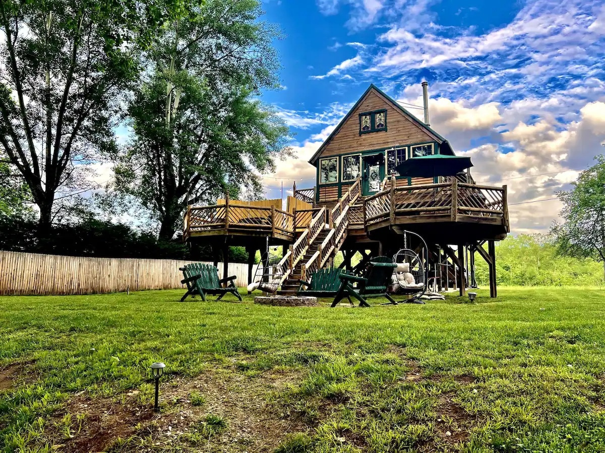 A spacious treehouse cabin is set on elevated stilts, surrounded by greenery. Multiple wooden decks feature seating areas and a covered porch. A fire pit is located nearby, with two swings hanging underneath the structure and steps leading to the entrance.