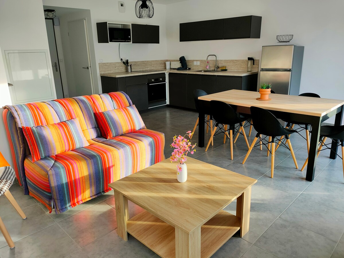 An open-concept living area features a cozy sofa adorned with colorful stripes, complemented by a wooden coffee table. A dining table with black chairs is positioned nearby, with a small potted plant providing a touch of greenery. A modern kitchen is visible in the background.