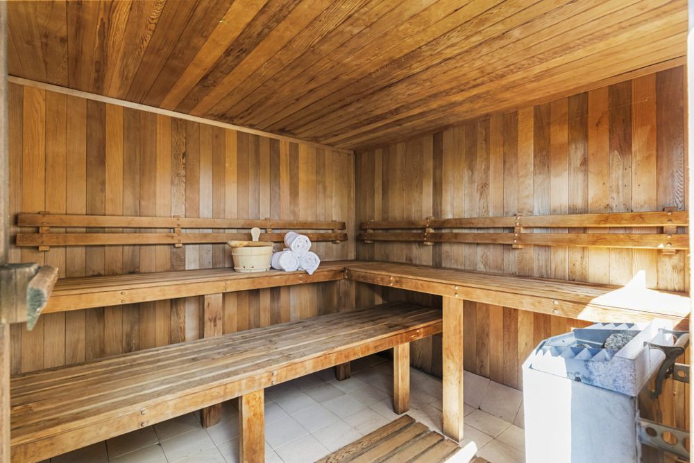 A traditional Swedish sauna is fitted with smooth wooden benches and vertical wood paneling. A storage bucket and neatly rolled towels are positioned on the bench. A metal container for sauna stones can be seen in one corner, contributing to the sauna's calming environment.