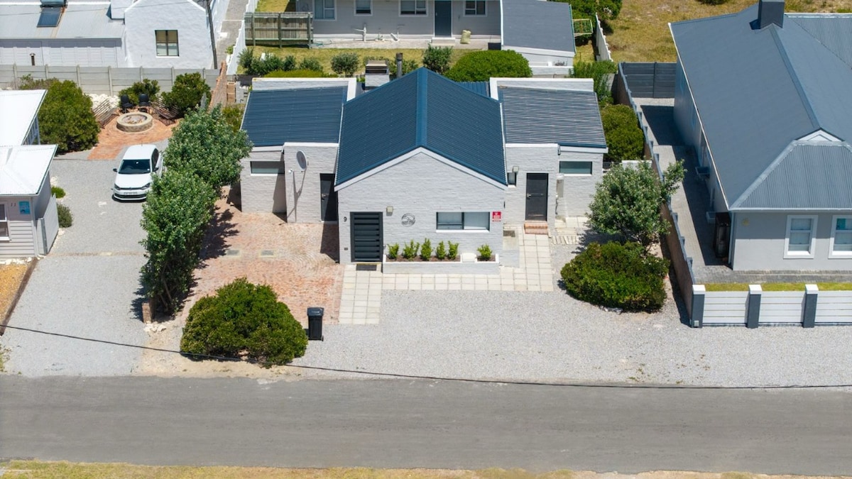 An aerial view reveals a modern house with a gray roof and light-colored exterior. Neatly arranged pathways lead to the entrance, surrounded by well-maintained shrubs. Nearby, a gravel driveway is visible, along with an outdoor seating area that features a fire pit.